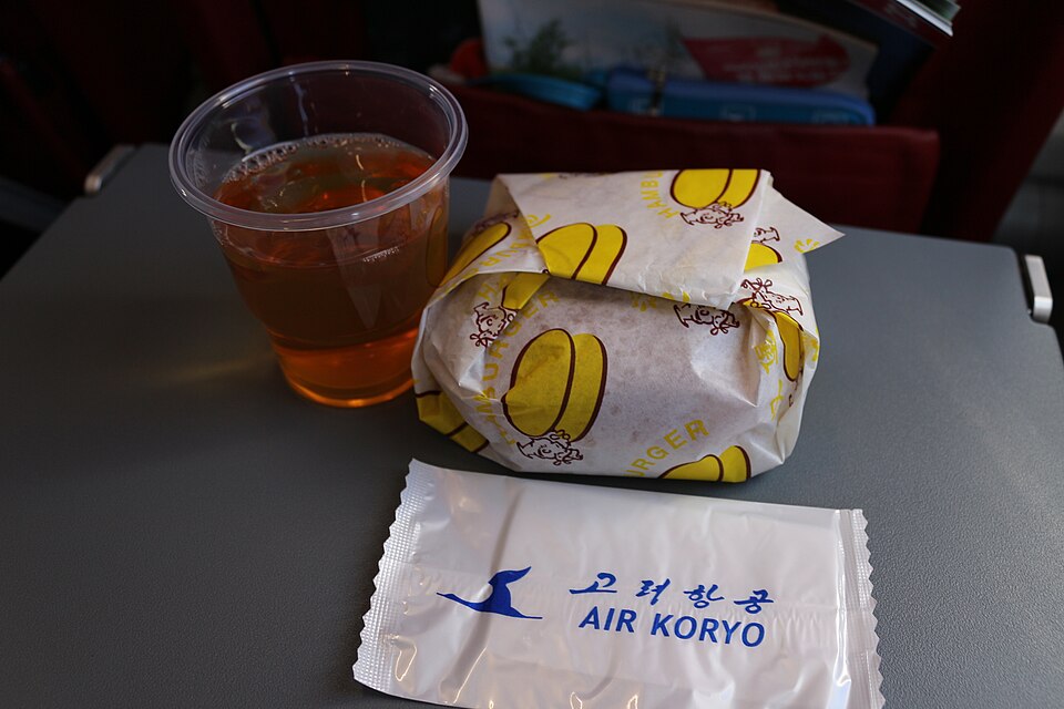 An in-flight meal on a tray table: a wrapped burger, a cup of juice and a napkin with an airline logo.