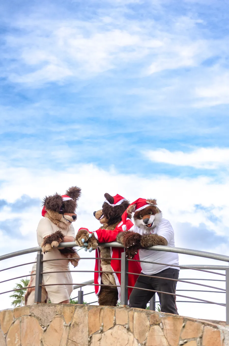 Three fursuiters leaning on a balcony railing under a blue sky.