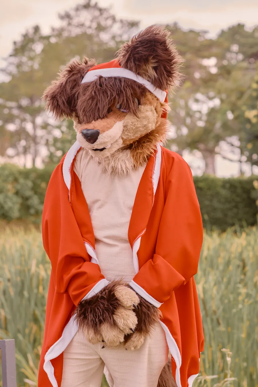 A fursuiter with a reddish fox-like head and red outfit standing in tall grass.