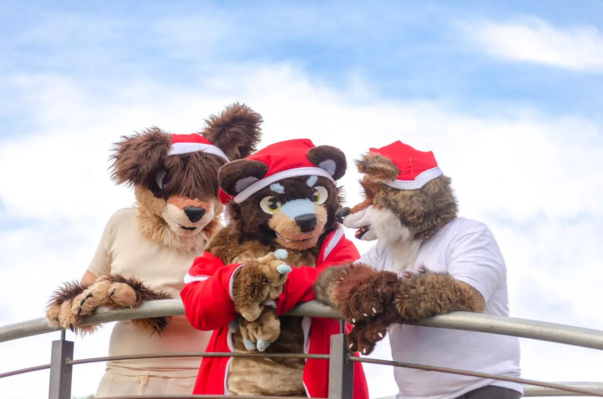 Three fursuiters in Santa hats leaning on a railing under a bright sky.
