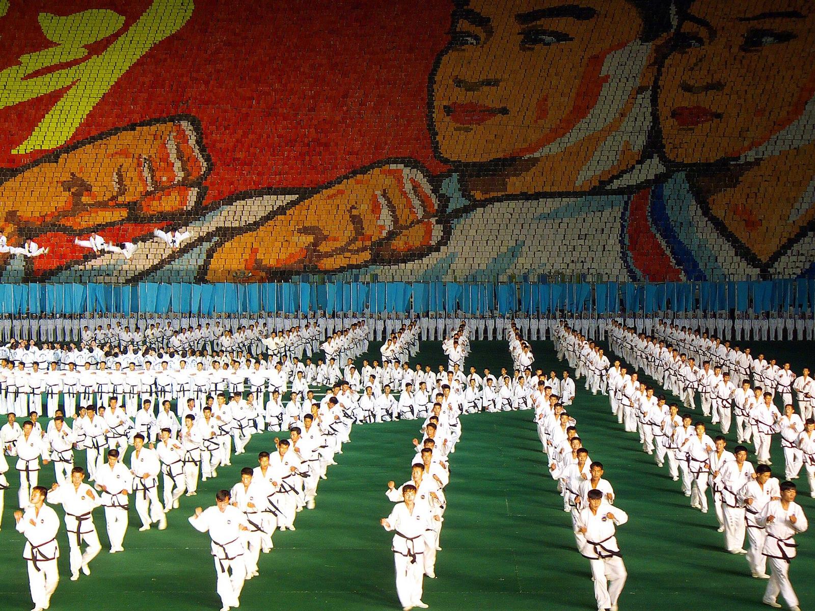 Thousands of performers in white on a stadium field during the Mass Games, beneath a giant card-stunt mosaic.