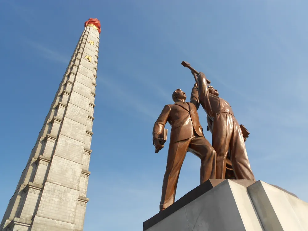 Looking up at the Juche Tower beside a bronze statue of three workers against a blue sky.