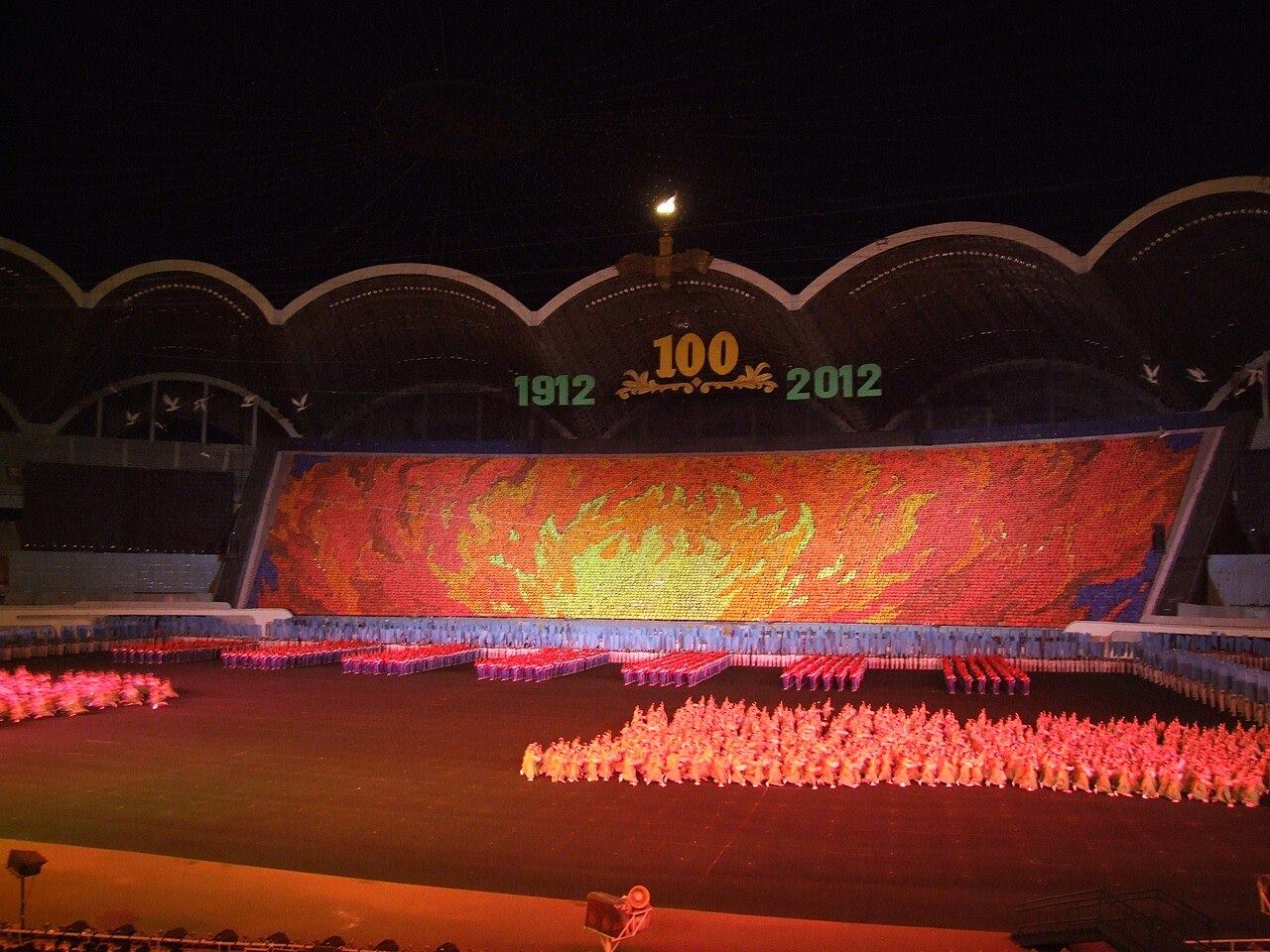 A packed stadium at night with a huge illuminated mosaic above a field of performers.
