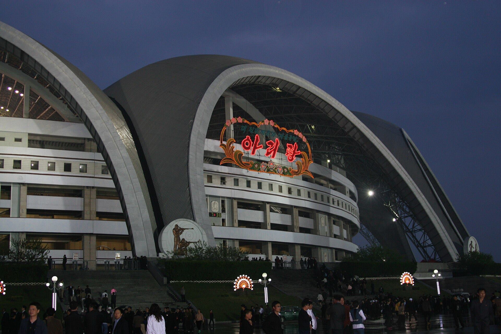 The scalloped white arches of May Day Stadium lit up at dusk, with a crowd outside.