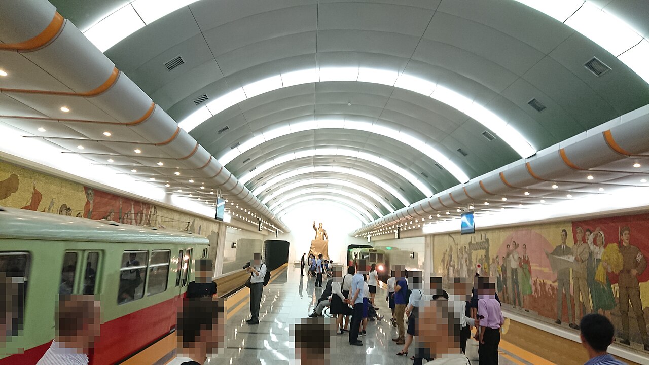 A long, brightly lit metro platform with an arched ceiling, a train on the left and murals on the right.