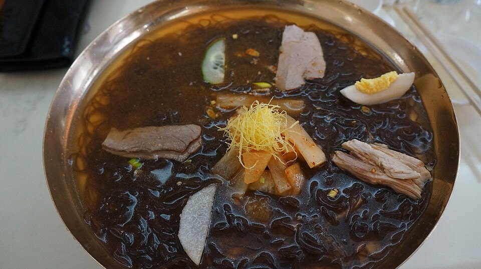 A bowl of cold buckwheat noodles in broth topped with sliced meat, egg and pear.