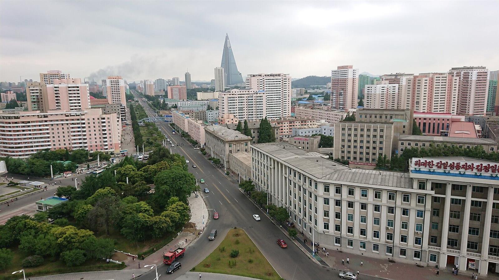 A wide avenue in Pyongyang lined with pastel apartment blocks, with the Ryugyong Hotel pyramid on the horizon.