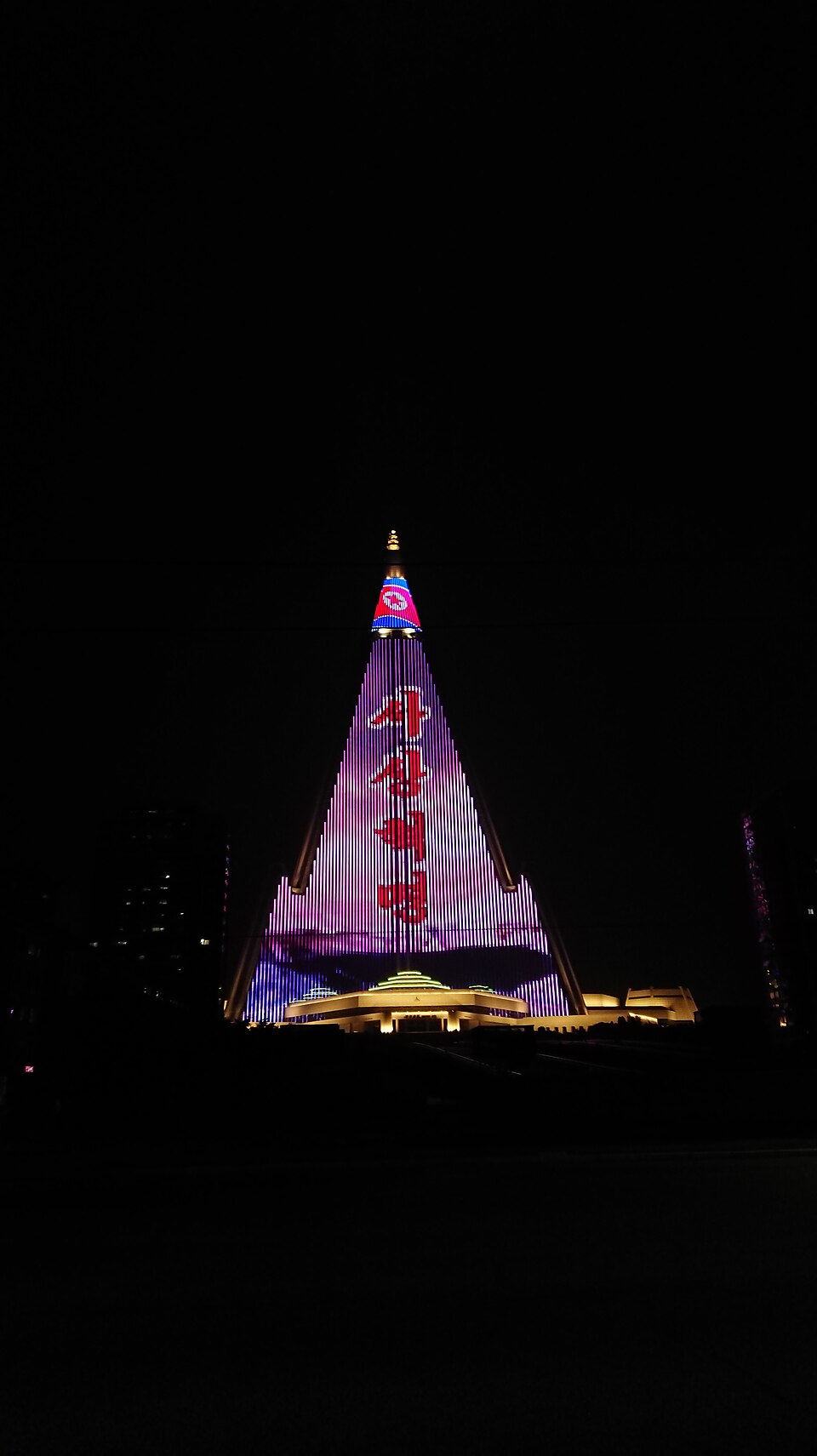 The pyramid-shaped Ryugyong Hotel lit up in pink and blue lights against a black night sky.