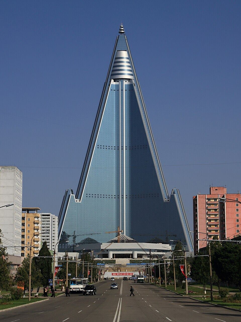 The Ryugyong Hotel's glass pyramid rising above an empty street and low apartment blocks under a blue sky.