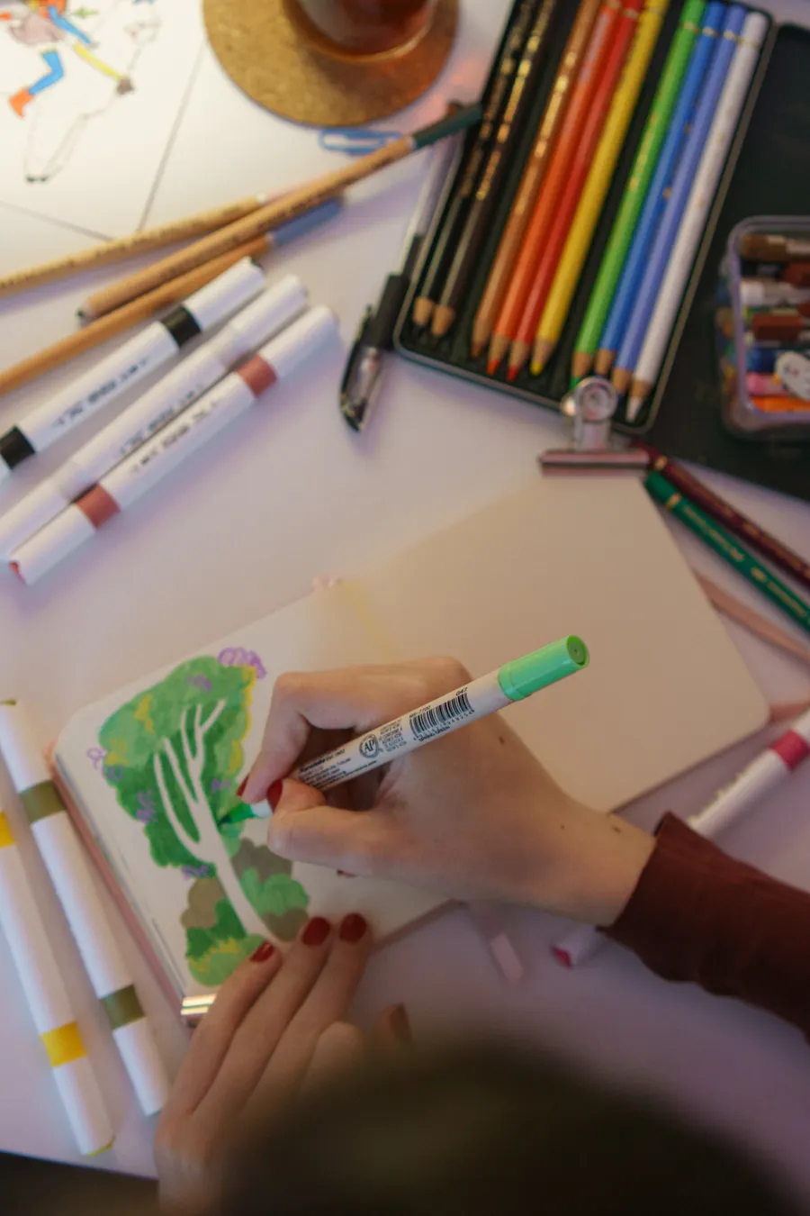 An artist drawing with markers in a sketchbook surrounded by pens and pencils.