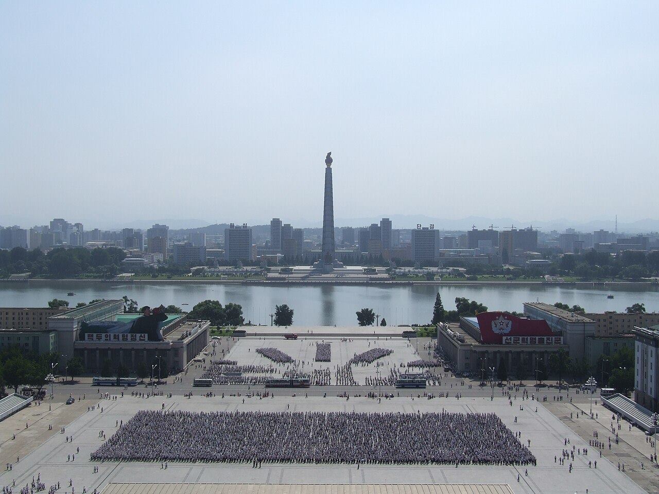 An aerial view across a huge square filled with rows of people in formation, the Juche Tower on the river beyond.